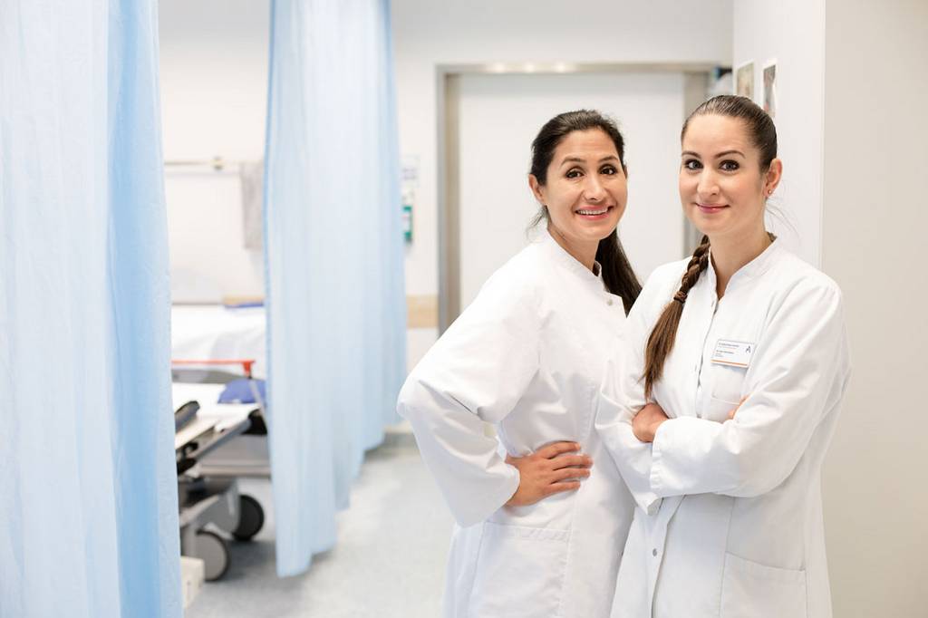 Zwei Ärztinnen, die in einem Krankenhausflur nebeneinander stehen und lächelnd in die Kamera schauen.